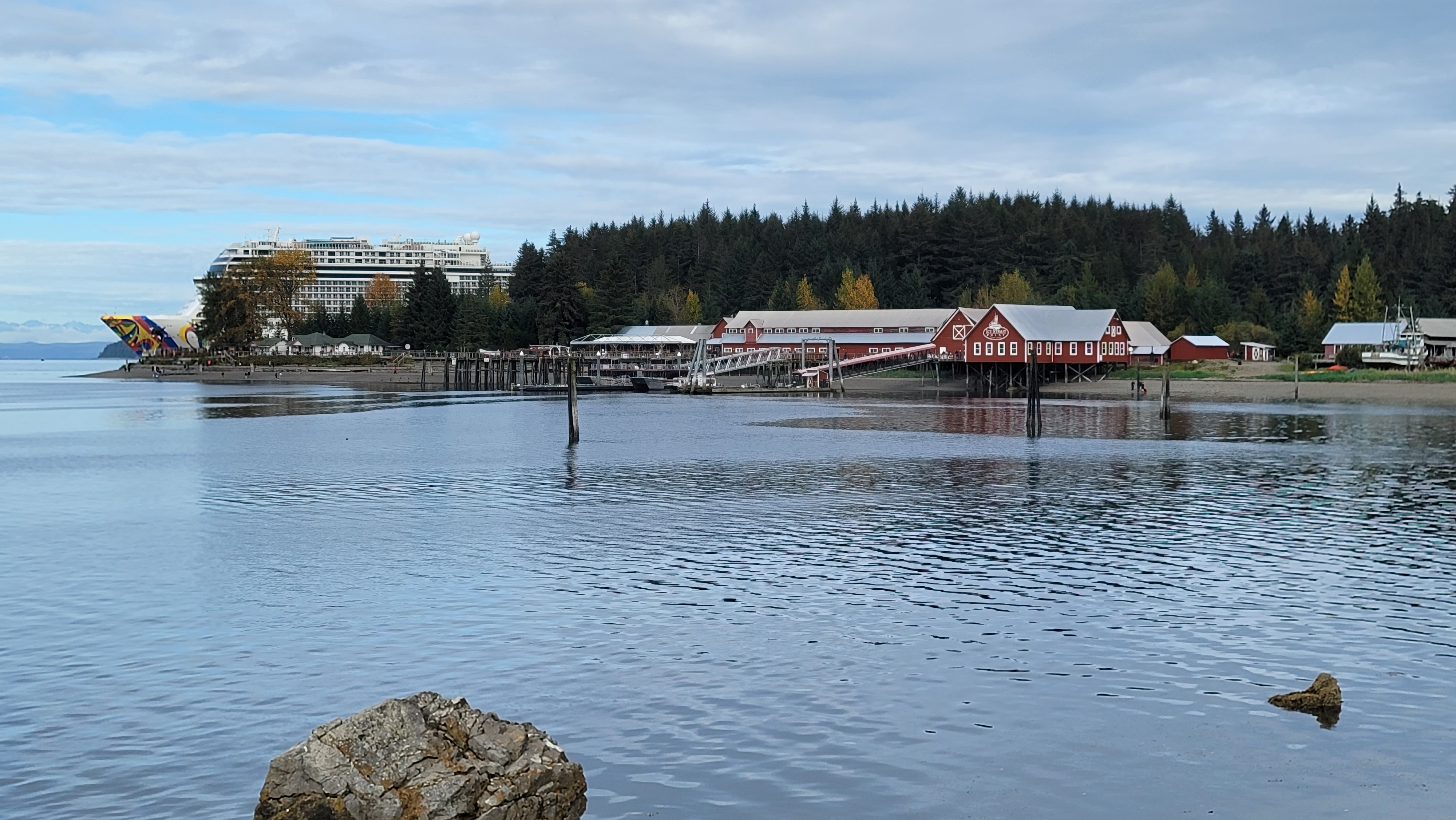 Red waterfront buildings at Icy Strait Point with forest and a cruise ship behind them.