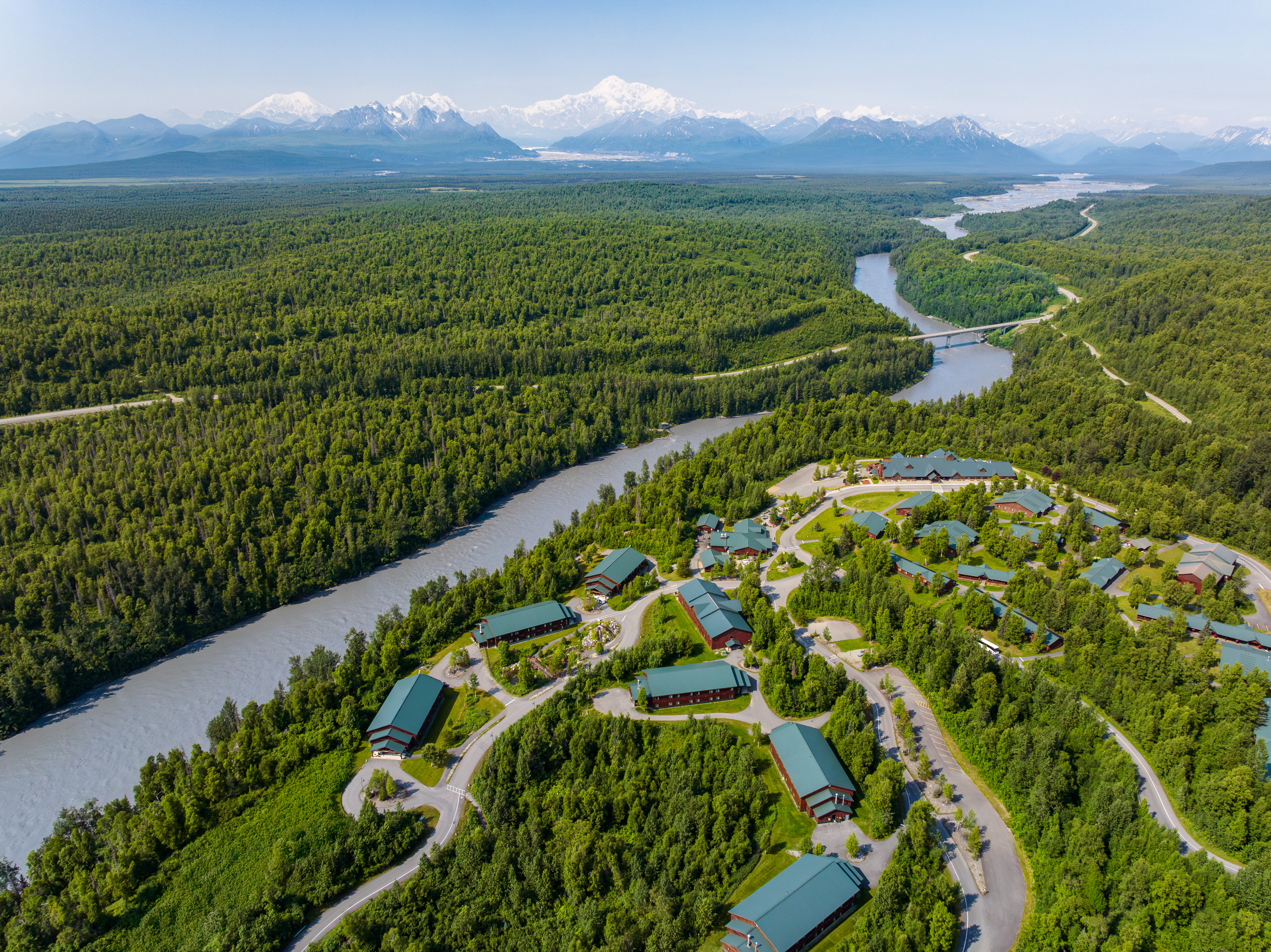 Aerial view of the McKinley Princess lodge grounds beside a river, with mountains in the distance.